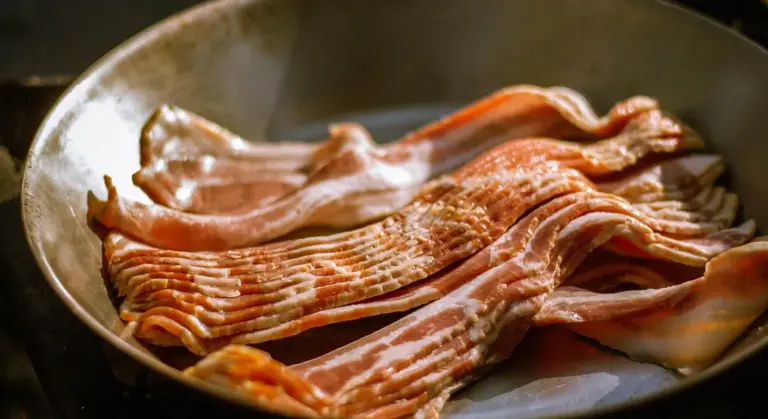 Thin strips of bacon frying in a cast iron skillet on a stovetop.