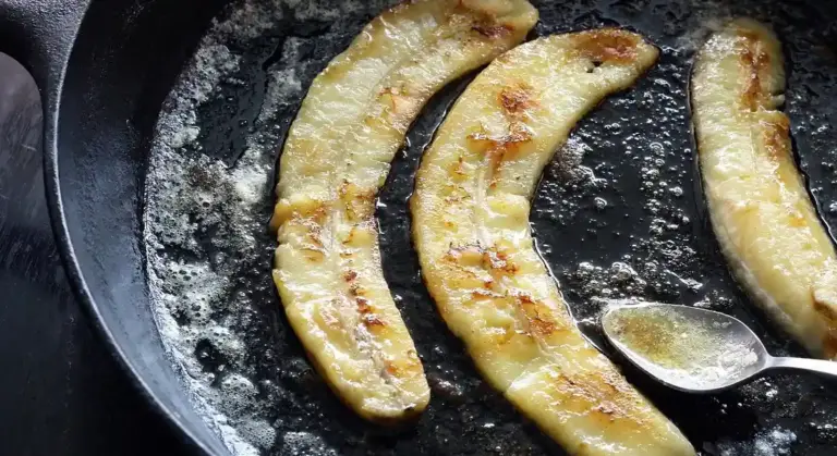 Banana slices browning in a cast-iron skillet with a small spoon resting in the pan.
