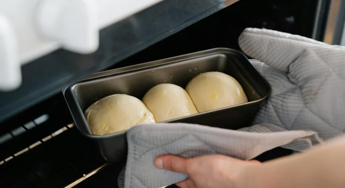 Three dough balls in a small loaf pan being placed into an oven, with gloved hands in quilted oven mitts.