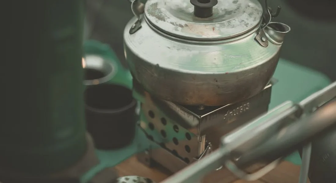 Close-up of a compact camping stove with a metal kettle on top, used for cast iron cooking over a campfire.