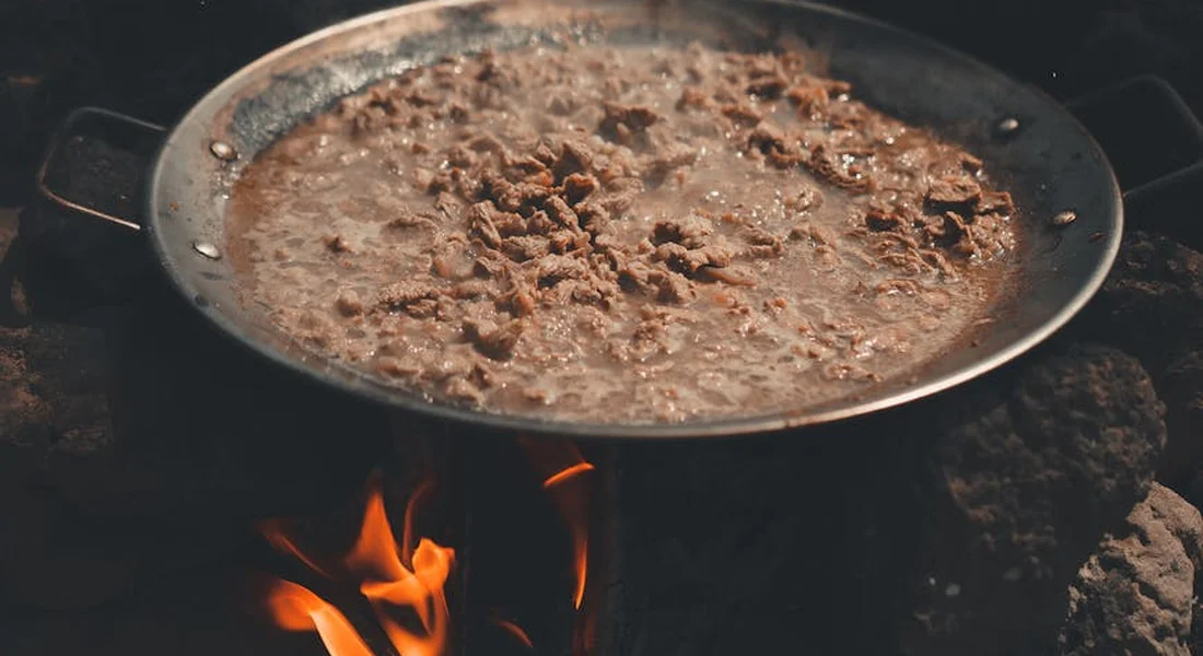 Cast iron skillet on a campfire with bubbling meat and orange flames underneath