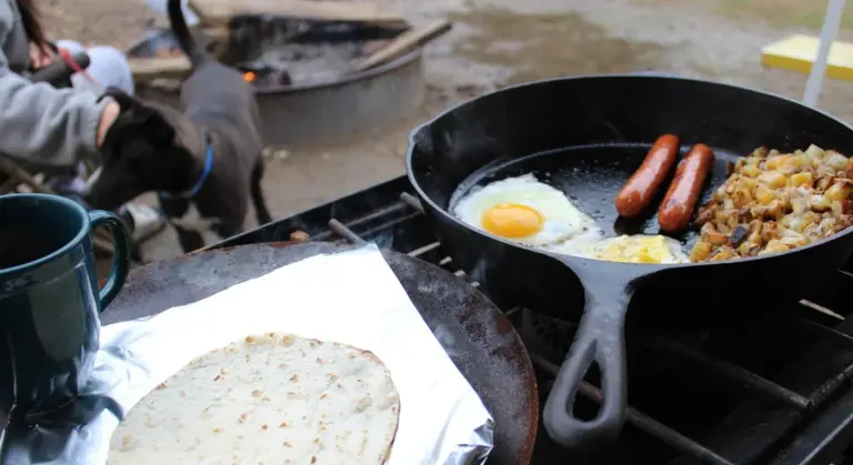 Cast iron skillet cooking eggs and sausages over a campfire with a tortilla on foil nearby.
