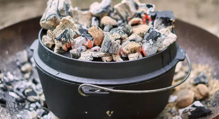 A black cast-iron pot with a metal handle sits on the ground, surrounded by hot charcoal and ashes.