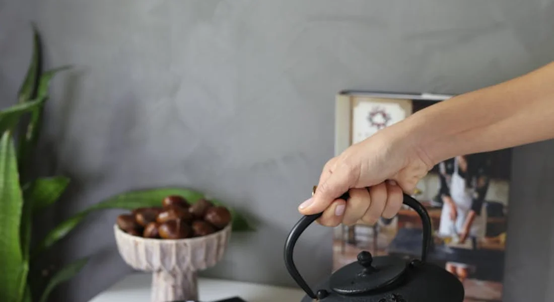 Close-up of a hand gripping the handle of a cast-iron kettle, with a bowl of nuts and a framed photo in the background.