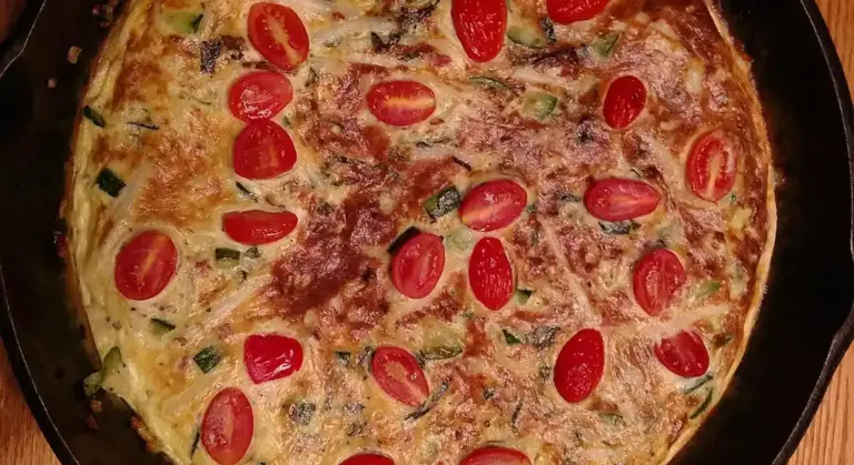 Cast-iron skillet on a wooden surface filled with a golden frittata, topped with cherry tomatoes.