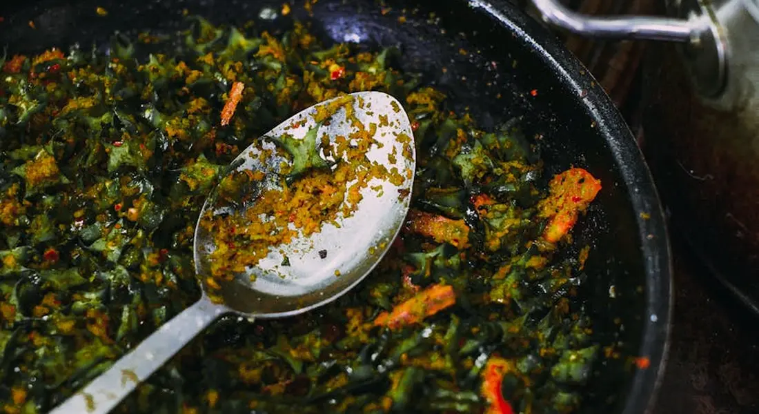 Close-up of a spoon resting in a skillet filled with sautéed greens and herbs.