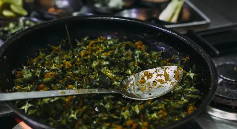 A carbon steel skillet containing sautéed greens with a metal spoon resting inside the pan.