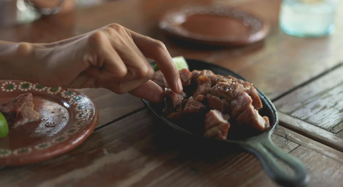 A hand lifts pieces of cooked meat from a cast iron skillet on a rustic wooden table.