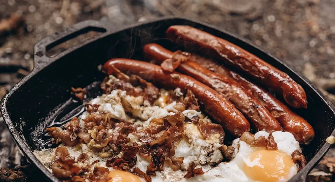 Cast iron skillet with sunny-side-up eggs and sausages cooking.