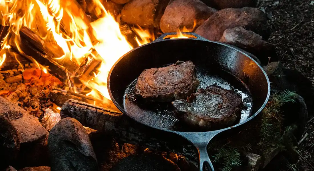 Cast-iron skillet with two chunks of meat sizzling over a bright campfire, surrounded by rocks.
