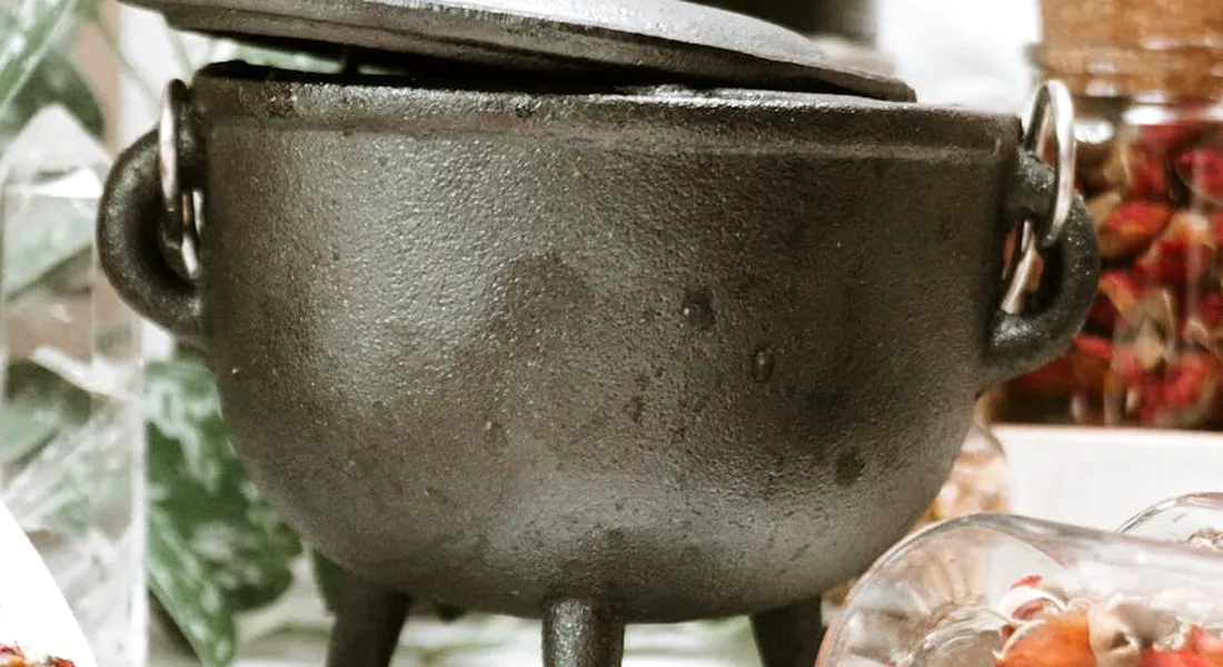 Cast iron pot with lid on a small charcoal grill, surrounded by herbs and kitchen items.