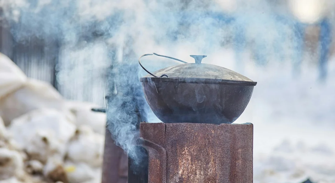 Cast iron pot on a rusty outdoor stove with steam rising, in a snowy outdoor setting.