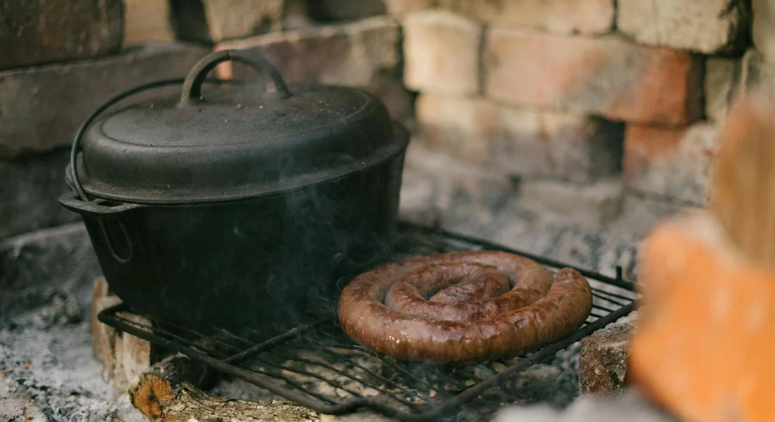 A cast iron pot with its lid rests on a grill over glowing coals in an outdoor setting, with a bread ring nearby.
