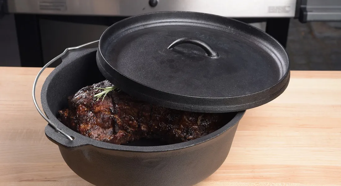 Cast iron Dutch oven with its lid slightly open, showing dark burnt-on residue inside; a small rosemary sprig rests on the surface.