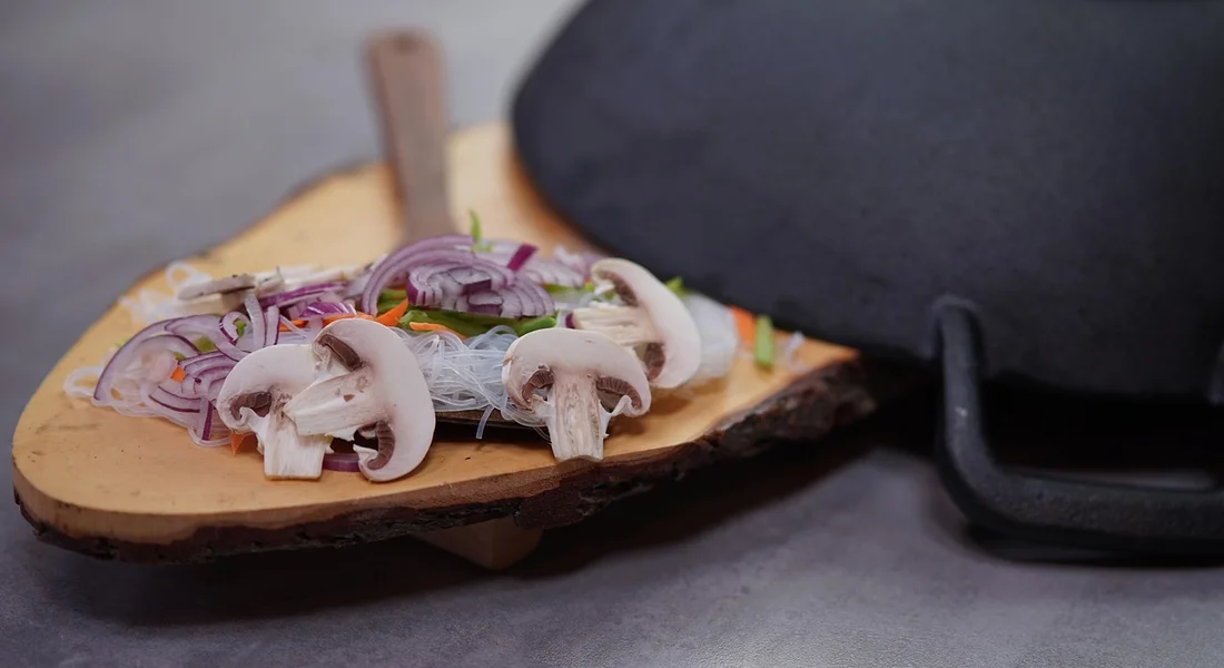 Mushrooms and red onion slices on a wooden cutting board with a dark bare cast-iron skillet in the background.