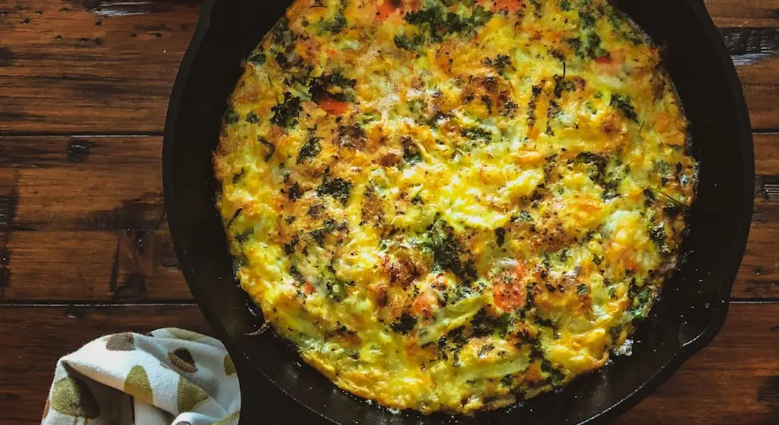 Golden baked frittata in a round black cast iron skillet on a wooden table with a folded cloth nearby.