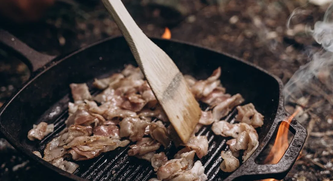 Cast-iron grill pan with a ridged surface cooking bite-sized meat; a wooden spatula rests across the pan as smoke rises.