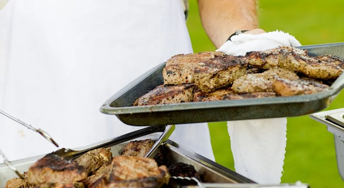 Outdoors, a cook lifts a tray of grilled meat from a barbecue while another tray sits nearby, showcasing well-seared pieces on cast-iron grill grates.