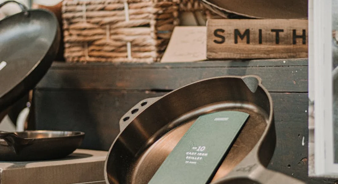 Cast iron skillets and pots arranged on a rustic shelf, with a wooden sign reading SMITH in the background.