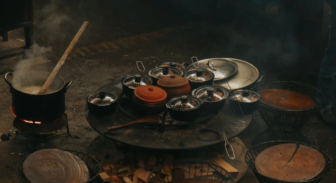 Open-hearth cooking scene with a circular grill holding several small cast iron pots, steam rising from a pot on the left as wood burns beneath.