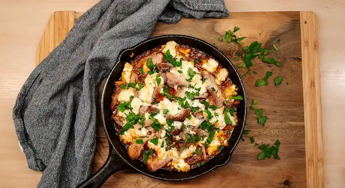 Cast iron skillet on a wooden board filled with a cheesy dish garnished with parsley, with a gray cloth nearby.