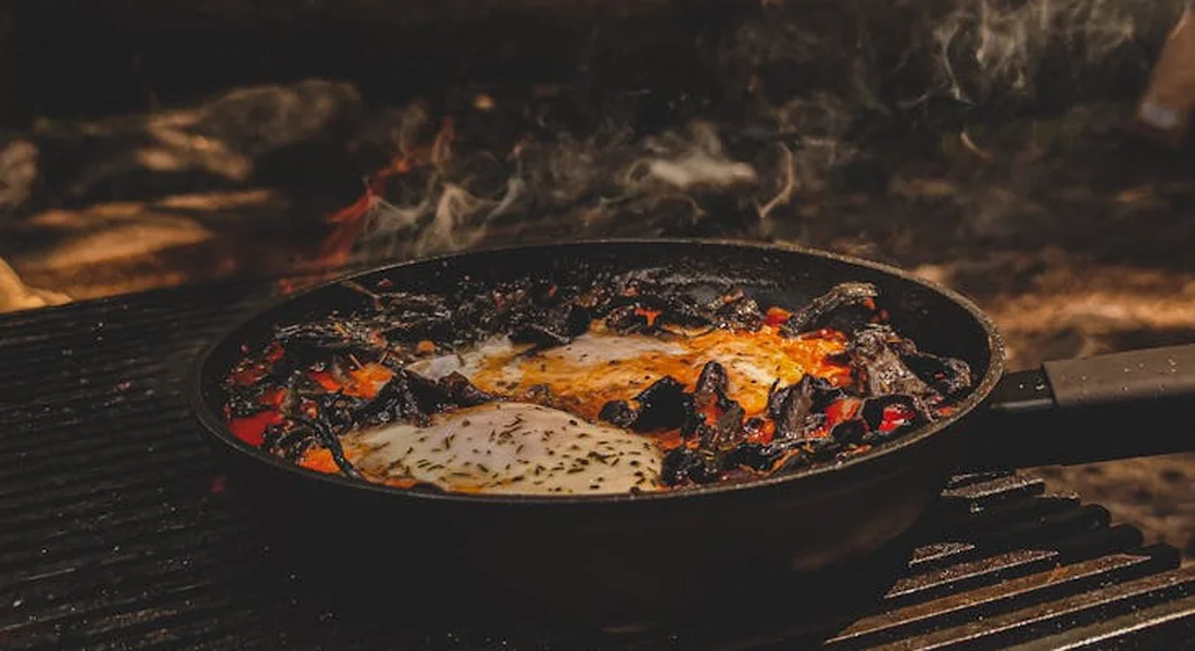 Cast iron skillet on a charcoal grill over an open flame, cooking with steam rising