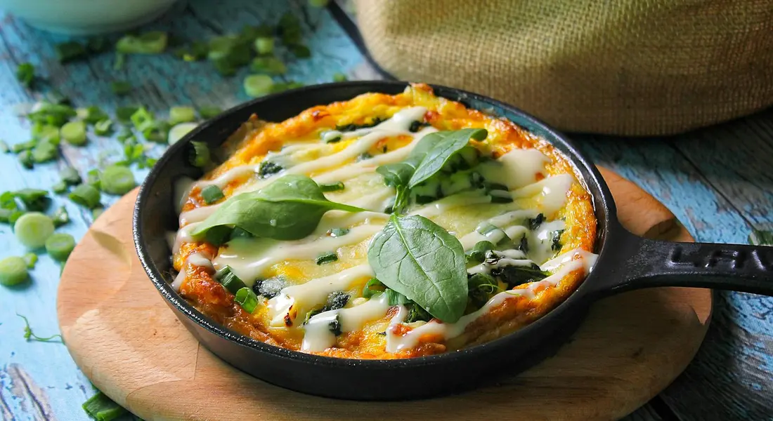 A savory pie or quiche baked in a small cast-iron skillet on a wooden board, topped with melted cheese and fresh green herbs.