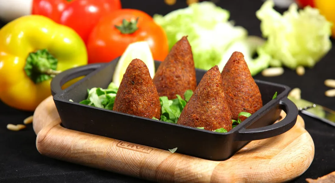 Cast iron skillet on a wooden board with falafel pieces, greens, and colorful peppers in the background
