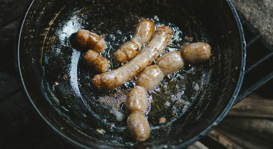 Sausages frying in a dark cast iron skillet with rendered oil and browned edges in a rustic kitchen.