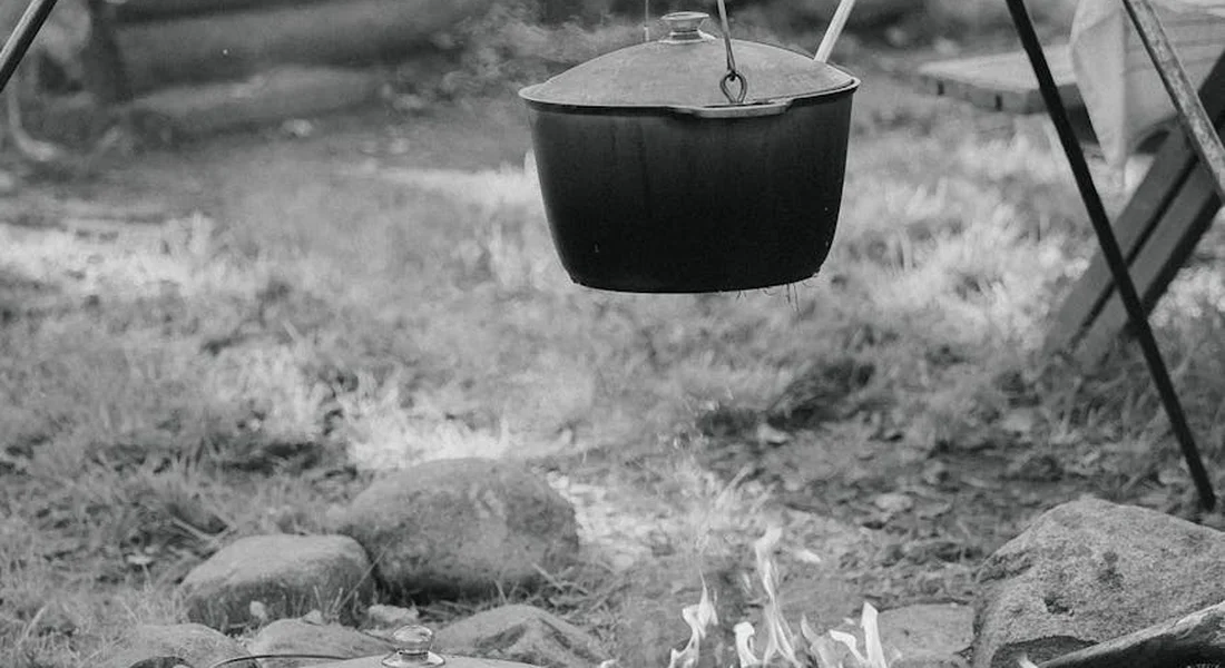 Black-and-white photo of a cast iron pot hanging from a tripod over a campfire, with rocks and dry grass in the background.