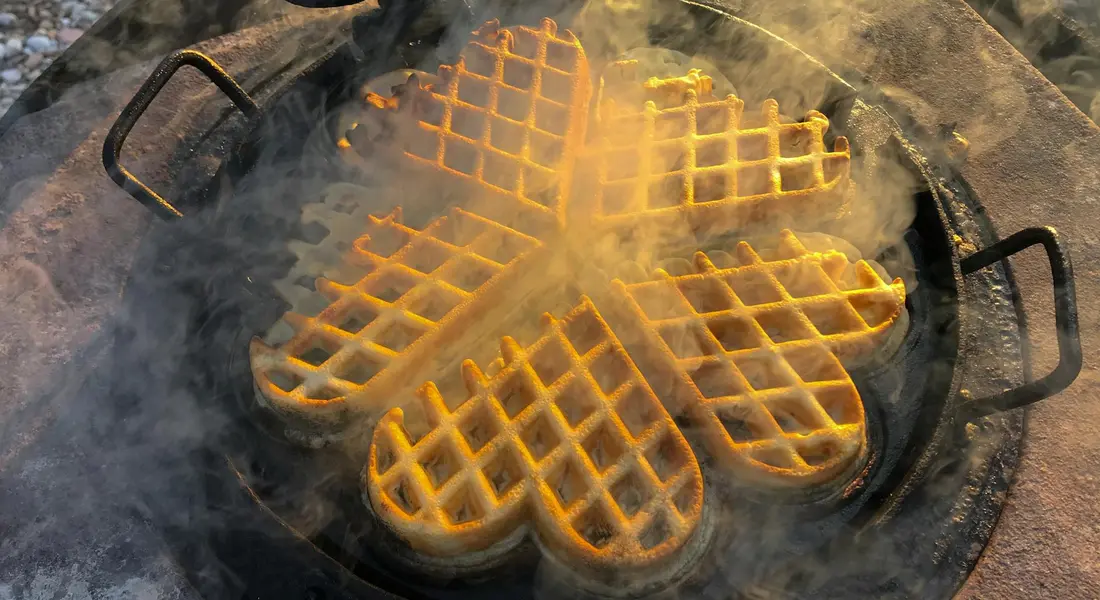 Outdoor scene of a round cast iron pan with four waffle-grid sections cooking; steam rises as waffles brown, illustrating why a well-seasoned surface matters for food release.