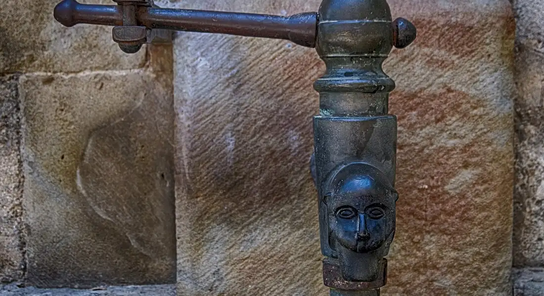 Close-up of a vintage metal faucet and pipe attached to a rough stone wall, showing weathered cast iron hardware.