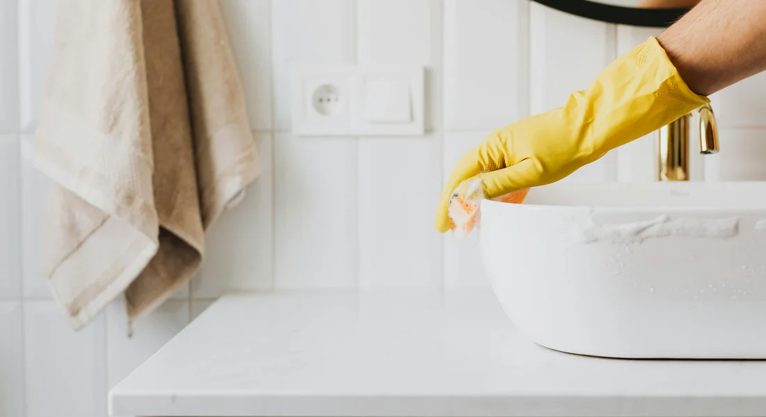 Gloved hand cleaning a white cast iron sink in a bright bathroom