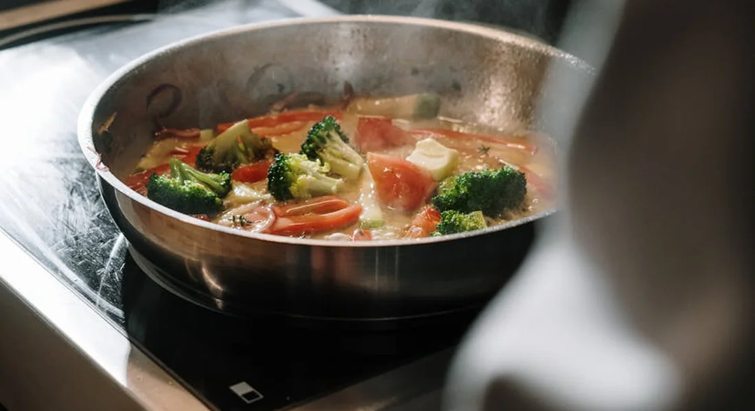 A cast iron skillet on a stovetop with bubbling vegetables like broccoli and tomatoes, steam rising from a simmering dish.