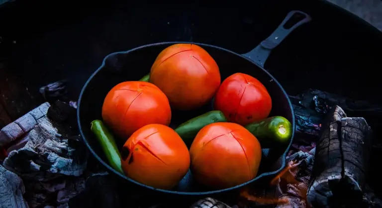 Cast iron skillet filled with ripe tomatoes and green peppers resting on outdoor leaves