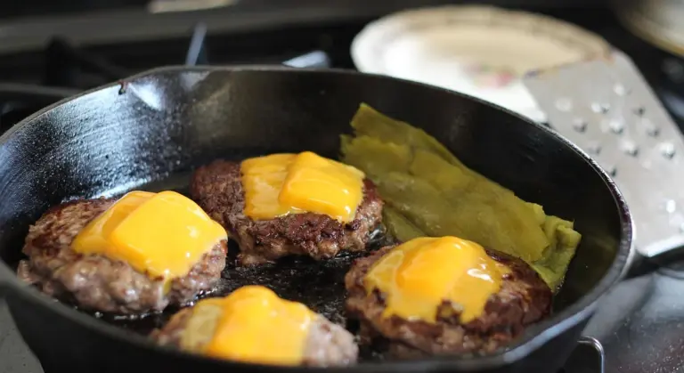 Cast iron skillet on a stovetop cooking several beef burger patties topped with melted cheese.