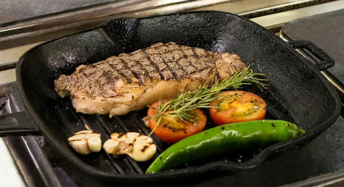 A steak sears in a cast-iron skillet on a stovetop, accompanied by roasted garlic cloves, cherry tomatoes, a sprig of rosemary, and a green chili pepper.