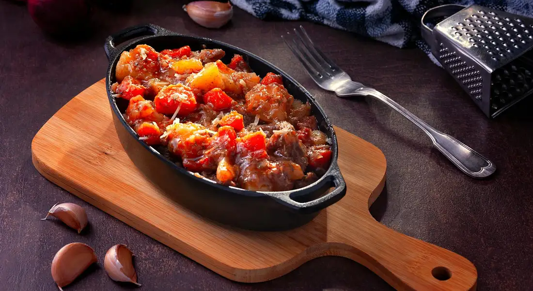 Cast iron skillet on a wooden cutting board filled with beef and vegetable stew, with a fork, a grater, and garlic cloves nearby.