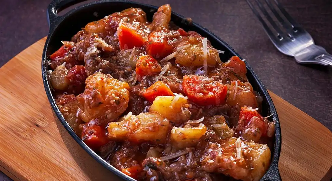 Cast iron skillet on a wooden board, filled with a chunky beef and vegetable stew, with a fork nearby.