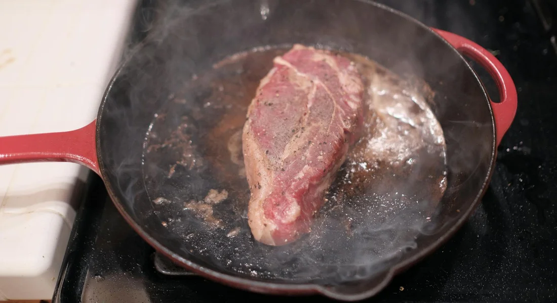 Cast iron tawa on a stovetop with a piece of meat searing in the pan, steam rising from the surface.