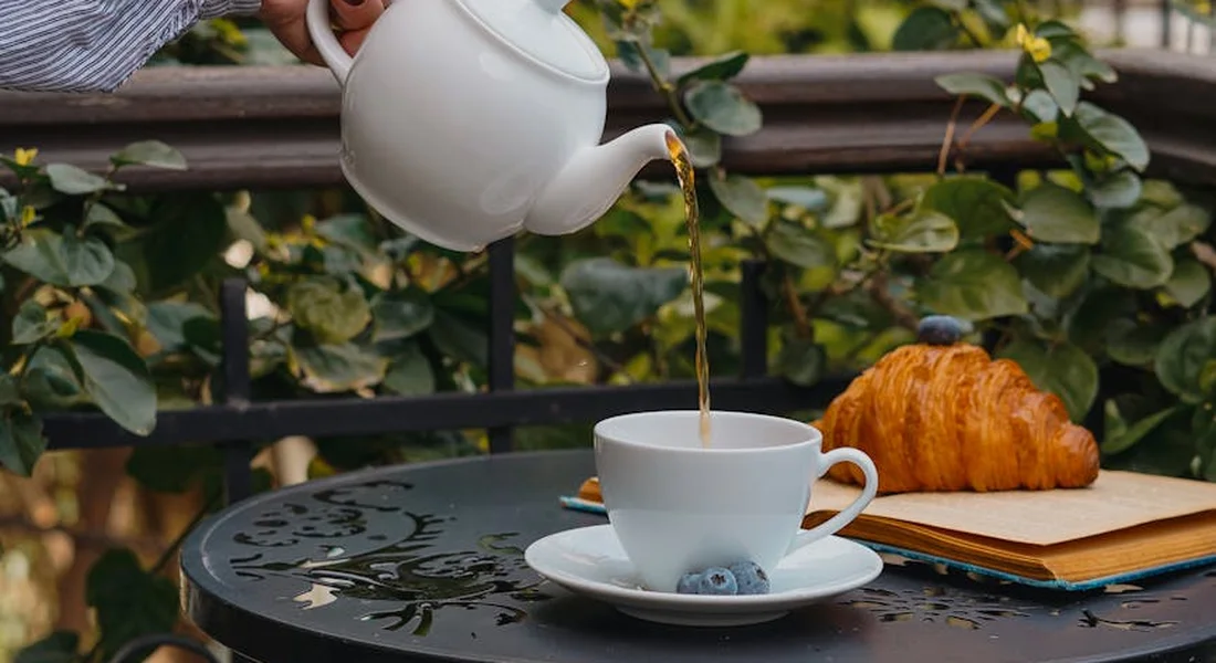 A white teapot pours hot water into a white teacup on a decorative outdoor table, with a croissant and notebook nearby.