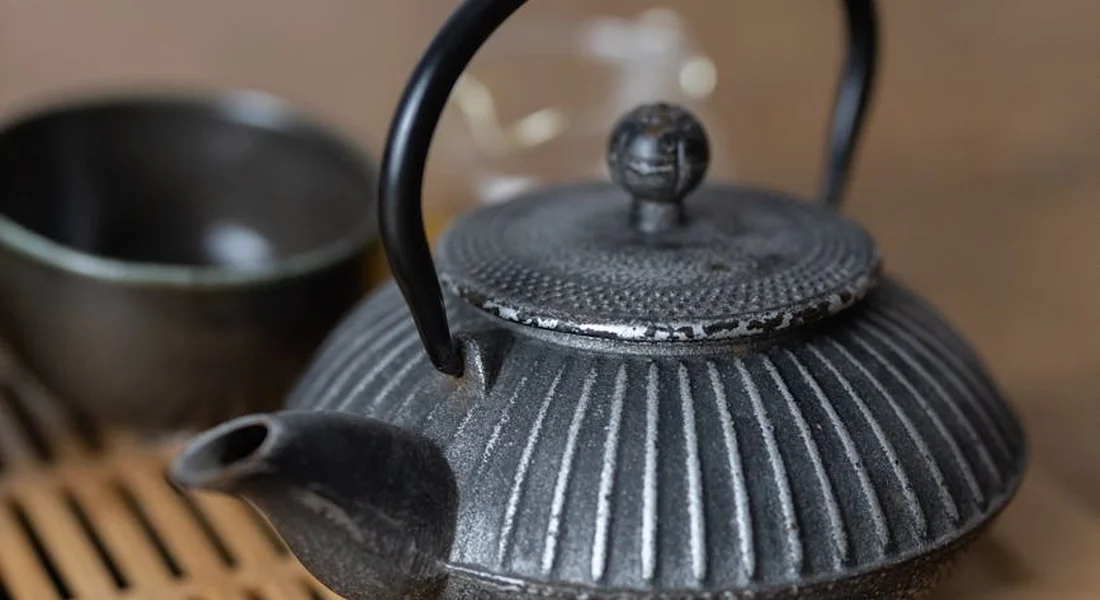 Close-up of a ribbed cast iron teapot with a curved black handle, sitting on a wooden tray with a blurred cup in the background.