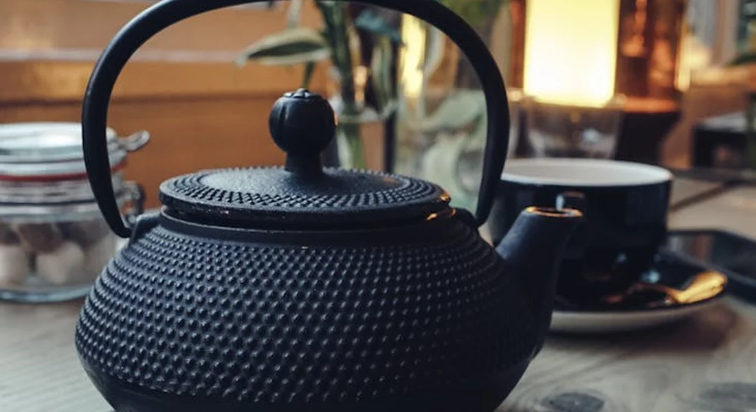 A textured black cast-iron teapot on a table with a cup and blurred kitchen background.