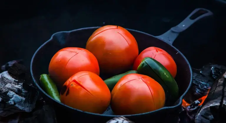 Cast iron skillet filled with orange tomatoes and a few green peppers resting on a rustic, dark surface.