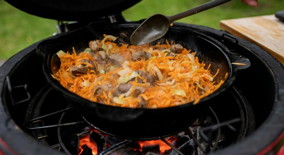 Cast iron skillet on a grill with a metal spatula stirring shredded vegetables (carrots) in the pan, flames visible underneath.