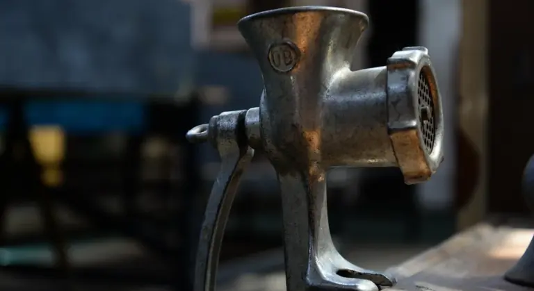 Close-up of a vintage metal meat grinder clamped to a surface with a worn dark finish.