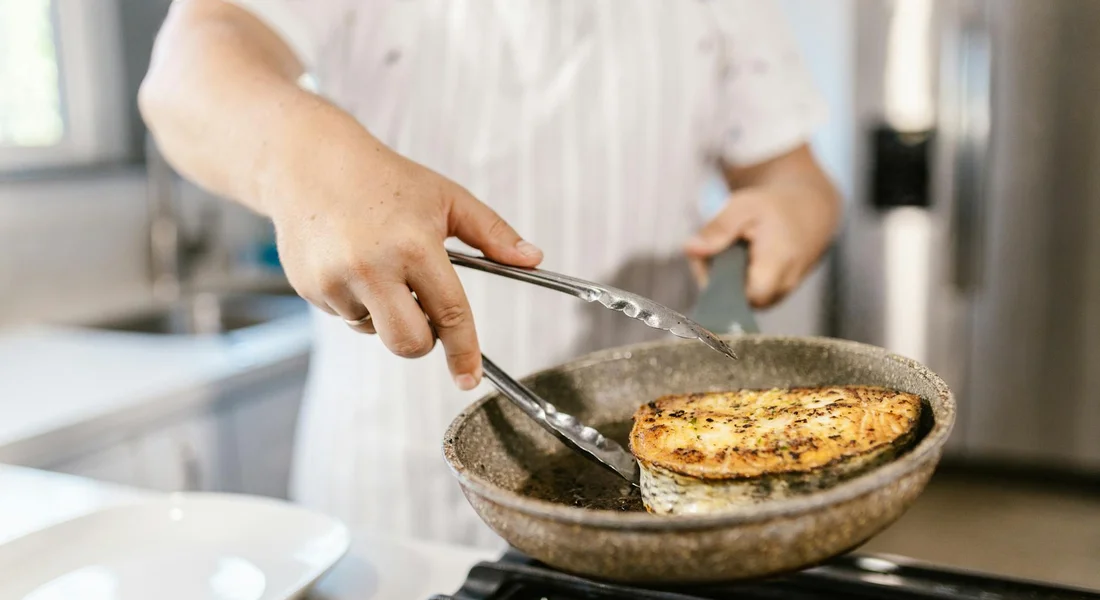 A cook in a white shirt flips a round, golden-brown patty in a dark cast iron skillet on a stovetop.