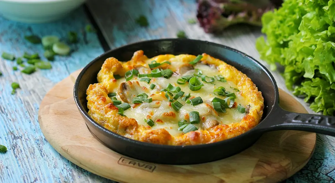 Cast iron skillet with a cheesy baked dish topped with green onions, on a wooden board with a blue tabletop.