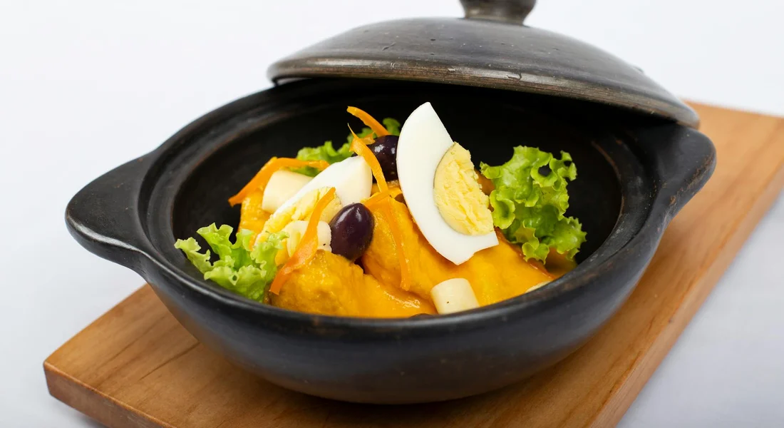 A black cast-iron pot with a lid slightly ajar sits on a wooden board, filled with a colorful dish garnished with greens and slices of egg.