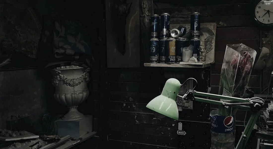 Dim workshop with a green adjustable lamp in the foreground, shelves of spray cans and a clock in the background, and a dusty workbench ready for stripping old cast iron seasoning.